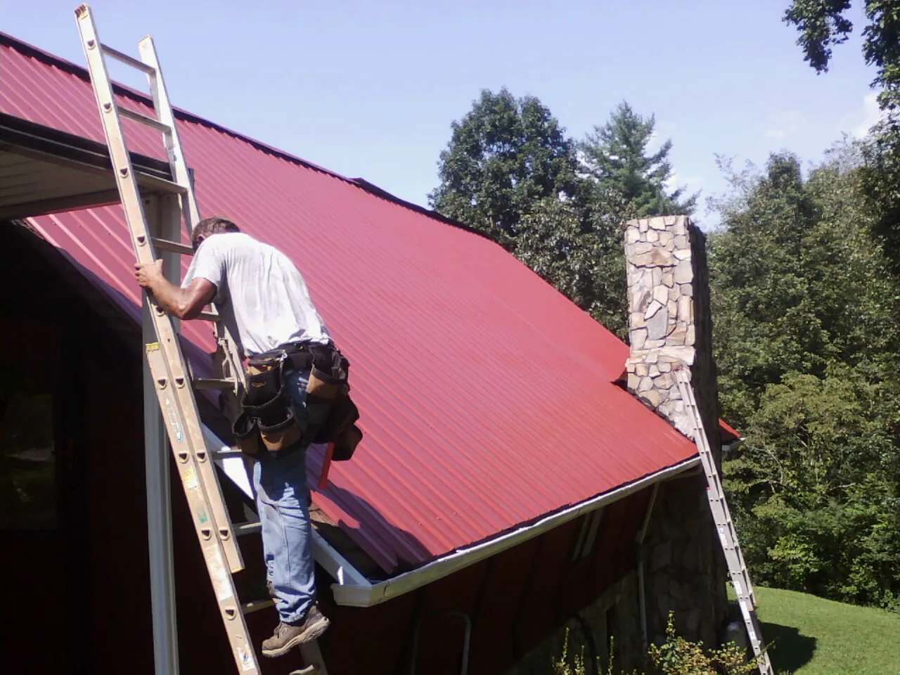 Roofer on a ladder beside a red metal roof and stone chimney — Soffit & Fascia Repair by Roofing Brothers in Onawa